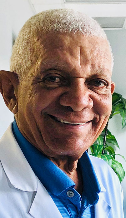 An elderly man in a blue shirt and white pants, smiling broadly, wearing glasses and a stethoscope, standing against a wall with a plant.