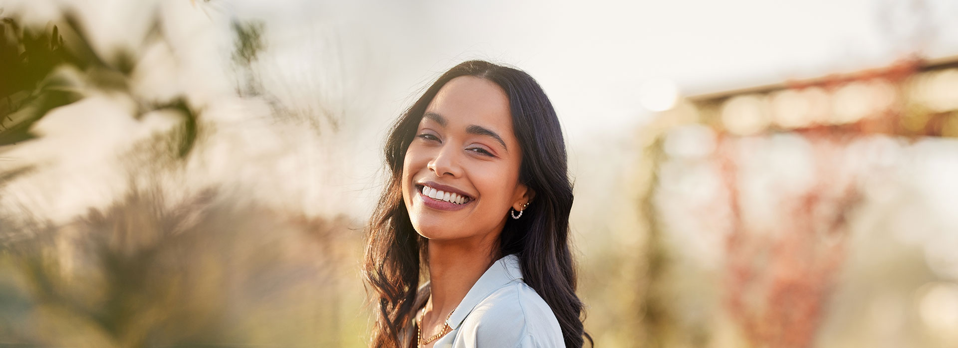 A smiling woman with long hair, wearing a white top, stands in front of a blurred background.