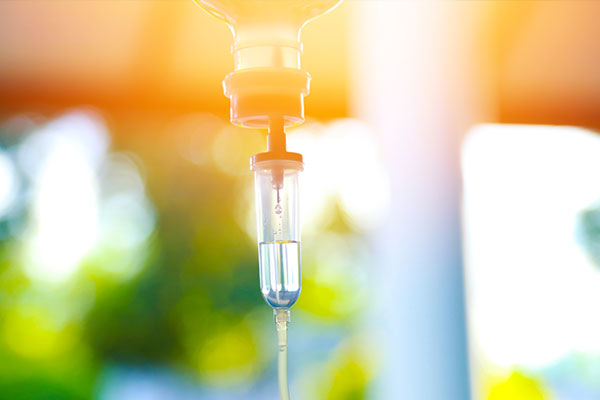 The image shows a clear glass syringe with a yellow liquid inside, attached to a drip line and connected to a metal faucet or spigot. The background is blurred but appears to be an outdoor setting with sunlight filtering through the leaves of trees.