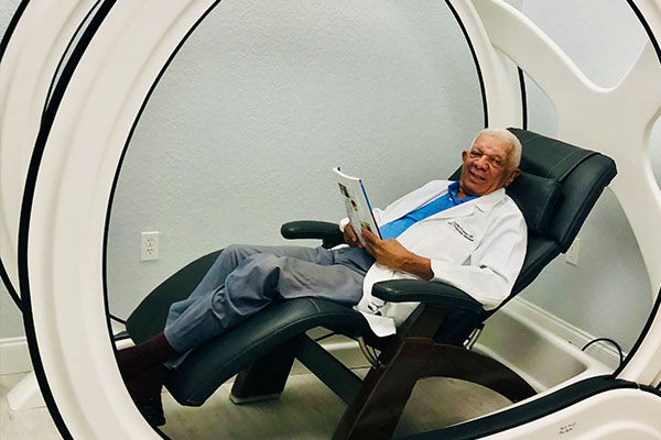 The image shows an elderly man sitting in a high-tech, circular chair with a futuristic design, wearing a white lab coat and holding a tablet.