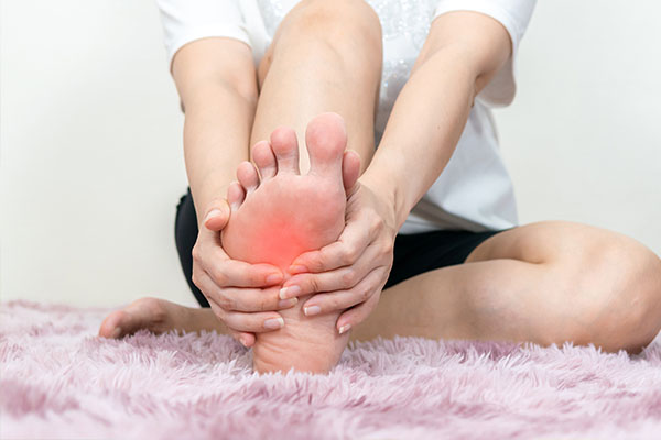 A person sitting cross-legged on a carpeted floor with their foot elevated, showing a visible red area, possibly an injury or inflammation, receiving apparent physical therapy.